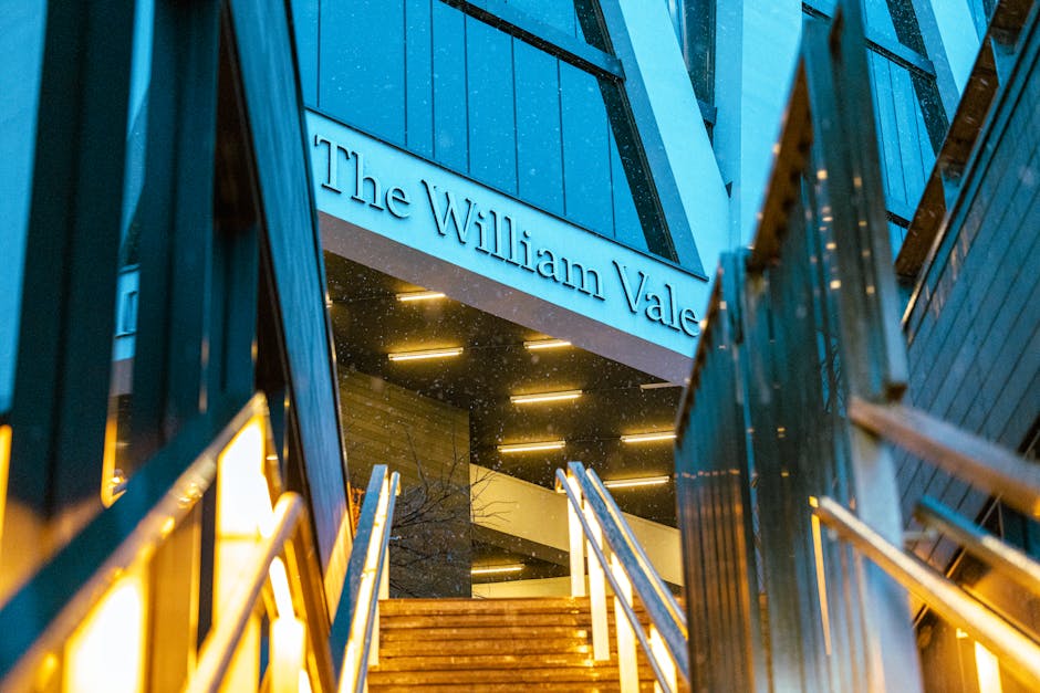 A modern multi-storey building with a prominent sign reading 'The William Vale' in metallic lettering mounted above a staircase leading to an entrance. The building's exterior features a combination of dark, textured materials and large glass windows, reflecting ambient lighting that illuminates the entrance area during twilight. The staircase has wooden steps with metal railings on both sides, guiding visitors upward. Surrounding the entrance are vertical metal and glass panels, adding to the contemporary architectural style. In the background, the building's facade displays horizontal lighting fixtures that enhance visibility and highlight the structure’s design. The scene captures a professional, urban environment suitable for private or corporate access, with a focus on the building's entrance as a focal point. This visual context aligns with concepts of commercial space access, highlighting the importance of maintaining clear pathways free of obstructions or rubbish, consistent with private waste disposal practices supported by services like Waste Removal Maida Vale.