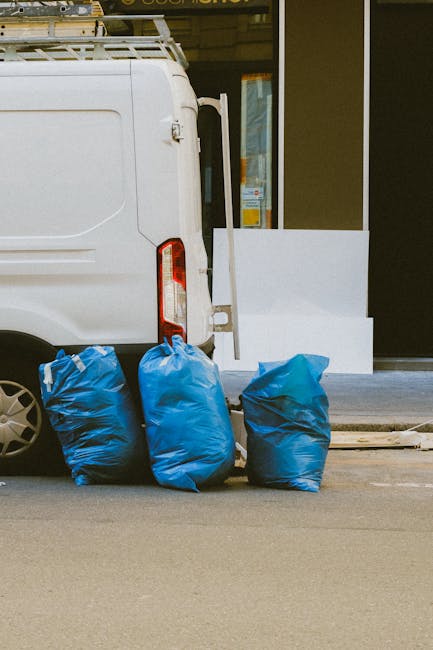 A white cargo van parked on a street with its rear doors closed, positioned beside a sidewalk in front of a commercial building with large windows and a dark entrance. In the foreground, three blue rubbish bags made of plastic are gathered on the pavement, filled with waste material, with a slightly crinkled texture and varying shades of blue. The bags are placed directly on the tarmac surface, which appears smooth and grey. Behind the bags, partially visible, is a wooden plank or a piece of debris lying flat on the ground. The van features a ladder rack on top, indicating sources of commercial or trades waste collection, and the environment suggests an urban or town setting where private rubbish removal, such as that provided by Waste Removal Maida Vale, is undertaken as an alternative to council services. The image captures a typical scene of on-site waste clearance, with natural light highlighting the textures of the bags and vehicle, supporting the context of professional rubbish removal services near Maida Vale.