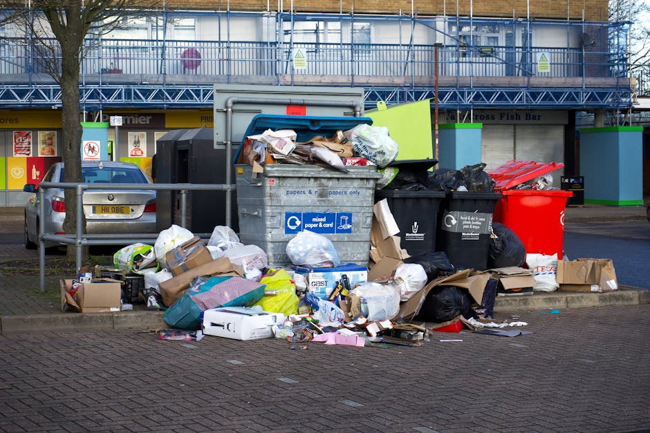 A large pile of mixed waste and rubbish overflowing from several black and red bins situated on a paved sidewalk, with various cardboard boxes, plastic bags, and paper scattered around the base of the bins. The debris includes crumpled papers, cardboard packaging, and plastic wrappers, some of which are spilling onto the street. Behind the bins, a grey metal container with a closed lid is visible, partially covered with a blue tarpaulin that is weighed down by additional rubbish. In the background, a building with a blue scaffolding facade and a row of commercial shopfronts, including signage for a fish bar, can be seen. A parked silver car is located to the left of the waste, and a metal railing separates the waste area from the vehicle. The scene is illuminated by natural daylight, highlighting the disorganized state of the discarded waste, which may reflect the need for alternative waste handling or rubbish removal services in the area.