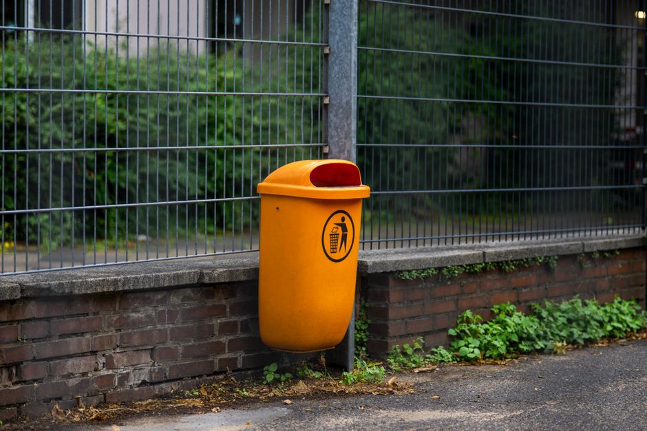 A bright orange outdoor waste bin is mounted on a concrete ledge below a metal fence, positioned on a paved sidewalk. The bin has a rounded, smooth surface with a contrasting red lid that features an integrated, slightly protruding handle for opening. On the front of the bin, there is a stick figure icon illustrating proper disposal into the bin, with an arrow pointing downward towards a smaller illustration of a trash can, indicating its purpose for rubbish collection. Behind the fenced area, lush green foliage with dense leaves and plants is visible, providing a natural backdrop. The concrete wall supporting the bin shows some moss and dirt accumulation, and small green plants or weeds grow at the base of the ledge, near the pavement. The overall scene appears clean and well-maintained, exemplifying a typical urban or suburban outdoor waste disposal setup that could relate to private waste handling or alternative rubbish removal services in a residential or community setting.