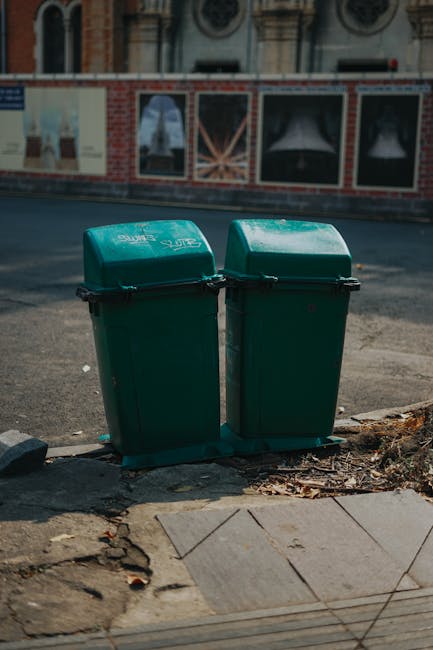Two green plastic rubbish bins with hinged lids are positioned side by side on a paved sidewalk, adjacent to a curb with some debris and fallen leaves. The bins appear to be made of durable, textured plastic with a matte finish and are marked with white text, likely indicating waste type or collection details. Behind the bins, there is an asphalt road with slight wear, and a brick and stone building facade featuring decorative window frames and ornate architectural elements, suggesting an urban environment. The scene is outdoors during daylight, with diffuse lighting that creates soft shadows, and the overall setting reflects a typical scene of local rubbish disposal, potentially related to private waste collection services offered by Waste Removal Maida Vale. The area shows signs of urban recycling or waste management, consistent with options for independent rubbish clearance in residential or commercial contexts in London.