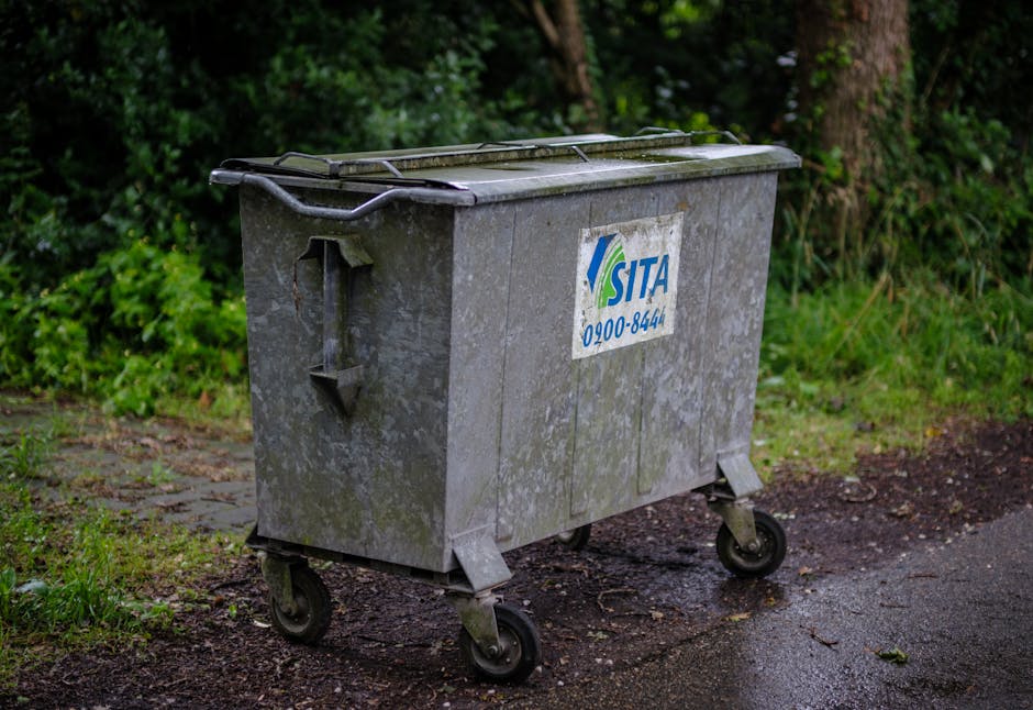A large, industrial metal wheeled bin with a rectangular shape, positioned on a damp asphalt surface adjacent to a grassy area with some patches of dirt and small plants. The bin's surface is weathered with visible streaks of dirt and rust, and it features a slightly domed, closed lid secured with metal handles on each side. A worn, rectangular white label with blue and green graphics, displaying the logo and contact number of SITA, is affixed to the front of the bin. The background includes dense green foliage and trees, suggesting an outdoor setting, possibly near a residential or commercial property. The overall scene is lit evenly, indicating it is daytime with natural diffuse light, which highlights the weathered texture of the bin and the surrounding natural environment. This image relates to the concept of private waste collection and rubbish removal services, like those offered by Waste Removal Maida Vale, which facilitate independent and on-site waste disposal options.