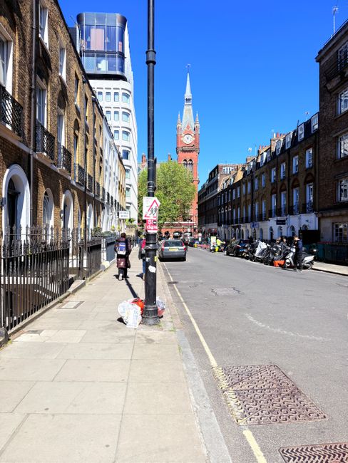 A wide city street scene in Maida Vale with a clear blue sky overhead. On the left side, there are brick residential buildings with iron railings and a sidewalk where a person is walking beside a large white plastic bag of rubbish. A black lamppost stands in the foreground, partially obstructing the view, with a small sign attached to it. Several parked motorcycles and motor scooters are lined along the right side of the street, with a few vehicles moving in the distance. In the background, a prominent red brick clock tower with a pointed spire is visible, rising above a mixture of modern and historic buildings, including the distinctive City Hall tower with its glass facade. The street scene includes elements typical of urban waste management, such as street-level rubbish bags, and the presence of Waste Removal Maida Vale occasionally visible in the context of independent waste collection or rubbish removal services.