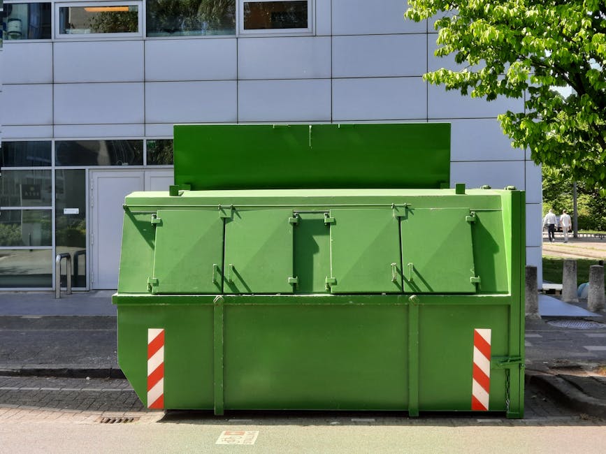 A large, industrial metal wheeled bin with a rectangular shape, positioned on a damp asphalt surface adjacent to a grassy area with some patches of dirt and small plants. The bin's surface is weathered with visible streaks of dirt and rust, and it features a slightly domed, closed lid secured with metal handles on each side. A worn, rectangular white label with blue and green graphics, displaying the logo and contact number of SITA, is affixed to the front of the bin. The background includes dense green foliage and trees, suggesting an outdoor setting, possibly near a residential or commercial property. The overall scene is lit evenly, indicating it is daytime with natural diffuse light, which highlights the weathered texture of the bin and the surrounding natural environment. This image relates to the concept of private waste collection and rubbish removal services, like those offered by Waste Removal Maida Vale, which facilitate independent and on-site waste disposal options.