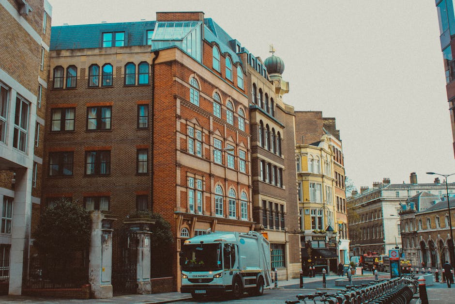 The image depicts a city street scene featuring a row of multi-story buildings with varied architectural styles, primarily constructed from red brick and stone. The prominent building in the foreground has arched and rectangular windows, with some topped by decorative architraves, and is complemented by a blue, metal-clad roof with multiple chimneys and small dormer windows. To the left, part of a modern white residential building with large windows is visible, contrasting with the older structures. On the street level, a blue and white waste collection truck with the branding 'Denmis' is parked near a gated entrance flanked by stone pillars and greenery, possibly indicating the premises of Waste Removal Maida Vale. Adjacent to the truck, there are no visible waste bins or rubbish piles, but the scene suggests an area designated for waste collection or clearance activities. The background features additional historic and commercial buildings, some with decorative facades, under an overcast sky which casts diffuse natural light across the scene. The overall setting combines urban architecture with waste management activity, illustrating practical aspects of private rubbish removal services in the area.