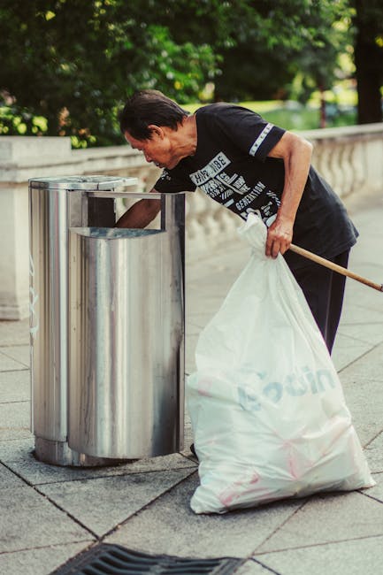A woman with short dark hair, wearing a black short-sleeved t-shirt with printed white text on the front, is seen emptying a large white plastic bag filled with rubbish into a stainless steel litter bin located outdoors on a paved walkway. The bin has a modern, rounded design with a vertical opening on top, positioned beside a low stone or concrete balustrade. In the background, lush green trees and a grassy area are visible, with sunlight filtering through the leaves, creating a bright and clear environment. The woman is slightly bent forward, holding the bag with her left hand while using her right hand to lift the lid of the bin, demonstrating a routine of trash disposal that could relate to private waste handling or independent rubbish collection services. The scene reflects a setting typical of urban or park environments where waste removal is managed discreetly, aligning subtly with the theme of rubbish removal services in Maida Vale.