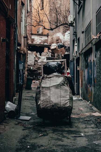 A narrow, urban alleyway cluttered with a large, wheeled bag made of worn, beige fabric in the foreground, surrounded by scattered debris and small items on the ground. Beyond the bag, heaps of cardboard boxes, plastic bags, and miscellaneous rubbish are piled up against an old brick wall at the back of the alley, which appears to be used for dumping waste. The alley is flanked by dark, graffiti-covered metal fencing on the left and a building with grey walls and barred windows on the right. Sparse, leafless tree branches are visible overhead, and parts of nearby buildings with pale, weathered facades can be seen in the background. The scene is lit with diffuse natural light, creating a somewhat bleak and cluttered atmosphere typical of a secondary waste collection site, which Waste Removal Maida Vale may handle as part of private rubbish management services.