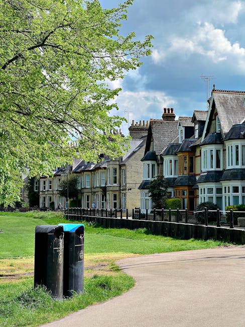 A row of traditional terraced houses with bay windows, painted in light pastel colors and featuring slate roofs, situated behind a black metal fence. In the foreground, a paved pathway runs alongside a grassy area with two black public rubbish bins placed near the edge of the grass. A large leafy tree with fresh green foliage extends its branches over the scene from the left, casting dappled shade. The sky above is partly cloudy with patches of blue visible. The environment suggests a residential street, where independent waste collection or private rubbish removal services, such as Waste Removal Maida Vale, might operate as an alternative to local authority waste disposal. The scene is calm and well-maintained, emphasizing the community-oriented nature of the area while subtly referencing waste management practices in a typical UK residential setting.