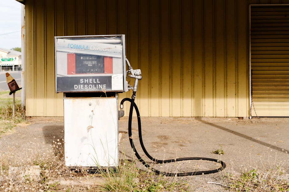 A vintage Shell gas pump with a white base and a rectangular display area at the top, featuring a faded metal facade with a protective glass cover, is situated outdoors on a concrete surface. The pump has a black rubber fuel hose coiled on the ground, connected to a metal nozzle attached to the right side of the pump. The background includes a weathered, vertical wooden wall painted in a muted yellow tone, with visible grain and some weathering. To the left, the edge of a driveway or pavement is visible, with a patch of grass and small weeds growing at the base of the pump. The scene appears to be in an open area, potentially part of a rural or less-trafficked property, with natural lighting indicating daytime. Waste Removal Maida Vale services may relate to the removal of older, unused fuel pumps or similar structures during site clearances as part of alternative waste handling or on-site clearance projects.