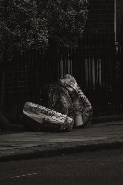 Two large black plastic bags of household rubbish, slightly crumpled and tied at the top, are placed on a grey pavement curbside in an outdoor setting. The bags appear to contain mixed waste materials and are positioned next to a black metal fence with vertical bars, behind which dense foliage from a tree or bush is visible. The scene is captured in ambient natural light, with shadows suggesting it may be late afternoon or early evening. The surrounding environment indicates an urban or suburban street context, possibly an area in need of rubbish removal services, such as those provided by Waste Removal Maida Vale, to support private waste handling and alternative collection options outside of local authority services.