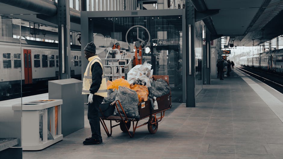 At a modern railway station platform, a worker wearing a high-visibility vest, dark clothing, a knit hat, and gloves stands beside a loaded trolley filled with various types of rubbish, including black and orange trash bags, cardboard, and plastic items. The trolley, positioned on the paved surface of the station, is in front of a glass-enclosed area with station machinery visible in the background. To the left, there is a white waste bin with an open top, and further to the right, the station platform extends towards a set of train tracks, where a few passengers are seen in the distance. The overall environment is illuminated by natural light, reflecting a clean and structured public space. The scene suggests a scenario of on-site waste collection, possibly handled by a private waste removal service such as Waste Removal Maida Vale, operating in a setting where independent rubbish disposal outside of local authority services is relevant, highlighting the logistical process of clearing waste from busy transportation hubs.