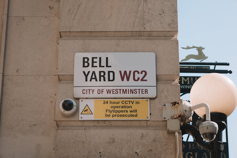 Close-up of a rectangular metal sign attached to a beige stone wall indicating a waste collection point at Bell Yard, WC2, managed by the City of Westminster. The sign features black and red text, detailing the location and waste type. Below the main sign, a smaller yellow notice with black text warns that CCTV is in operation 24 hours and that flytippers will be prosecuted. To the right of the signs, a white security camera is mounted, facing downward, alongside a decorative street lamp with a spherical, frosted glass cover and a black metal post. In the background, part of a street sign with a silhouette of a reindeer and some ornate black metalwork is visible against a clear sky. The scene depicts an urban area with a focus on designated waste disposal signage and security measures, consistent with private waste collection areas in Westminster.