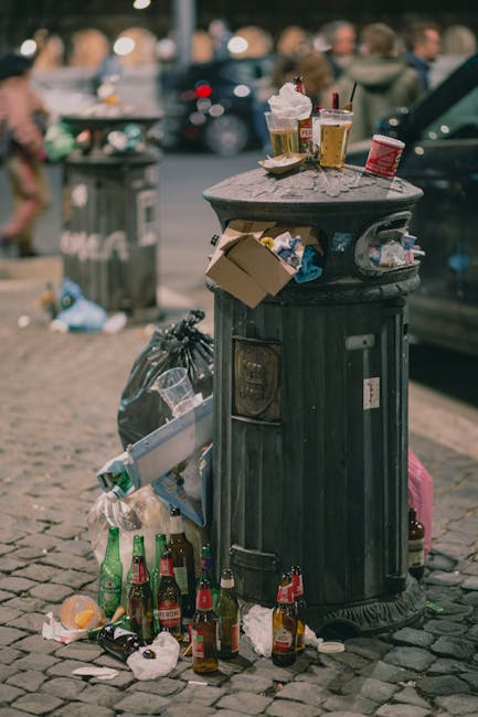Close-up of a rectangular metal sign attached to a beige stone wall indicating a waste collection point at Bell Yard, WC2, managed by the City of Westminster. The sign features black and red text, detailing the location and waste type. Below the main sign, a smaller yellow notice with black text warns that CCTV is in operation 24 hours and that flytippers will be prosecuted. To the right of the signs, a white security camera is mounted, facing downward, alongside a decorative street lamp with a spherical, frosted glass cover and a black metal post. In the background, part of a street sign with a silhouette of a reindeer and some ornate black metalwork is visible against a clear sky. The scene depicts an urban area with a focus on designated waste disposal signage and security measures, consistent with private waste collection areas in Westminster.